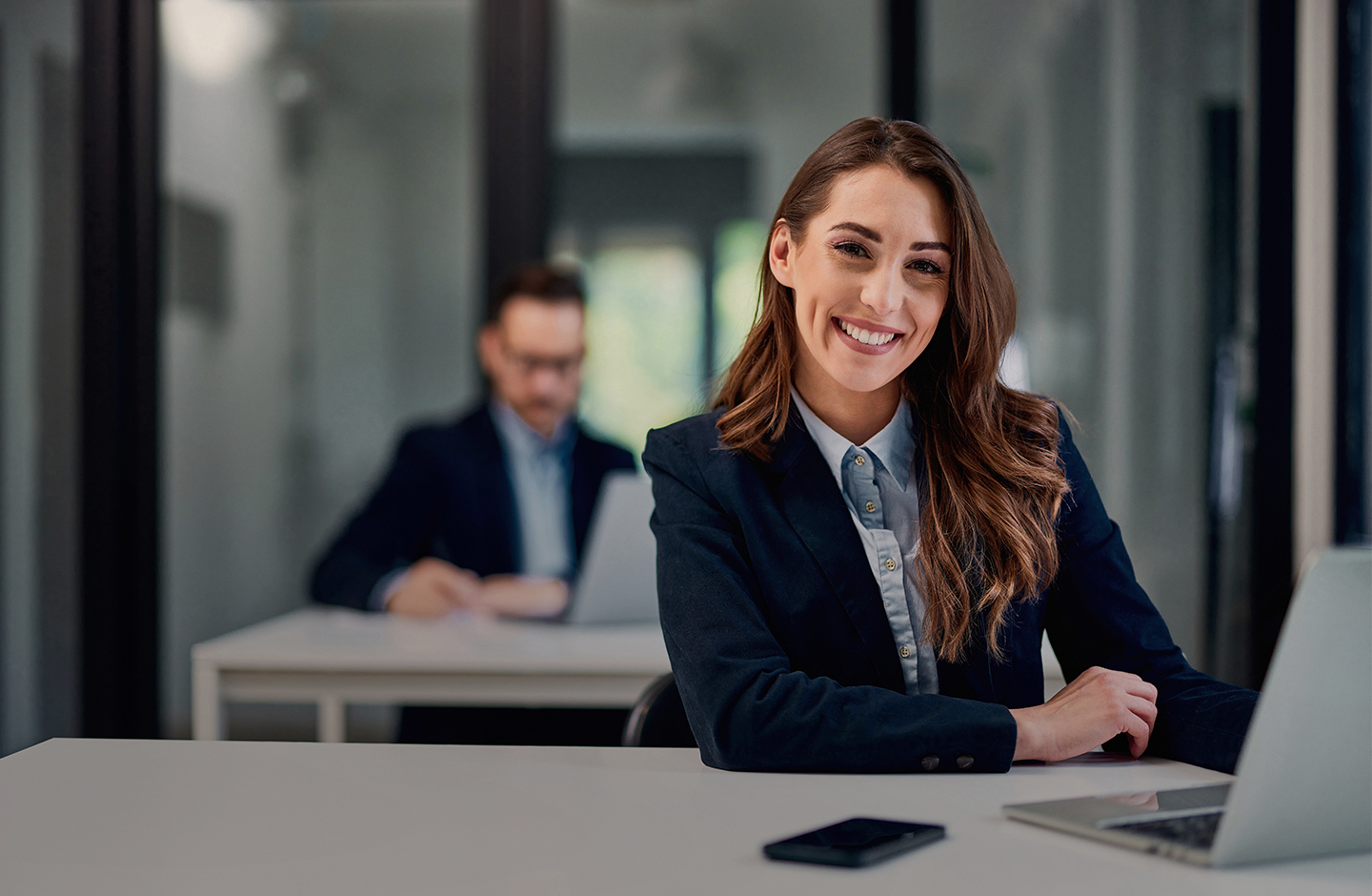 Eine Geschäftsfrau im Blazer sitzt lächelnd an einem Schreibttisch.