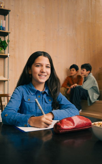 Ein Mädchen sitzt an einem Tisch bei den Hausaufgaben. Sie hält einen Stift in der Hand und lächelt in die Kamera.