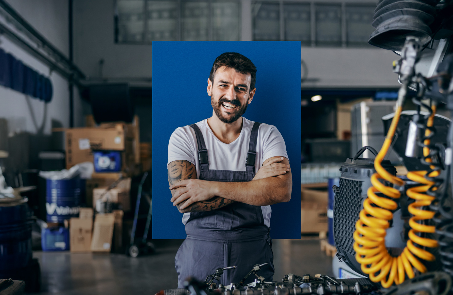Ein Mann in Latzhose steht in einer Werkstatt und verschränkt lachend die Arme.