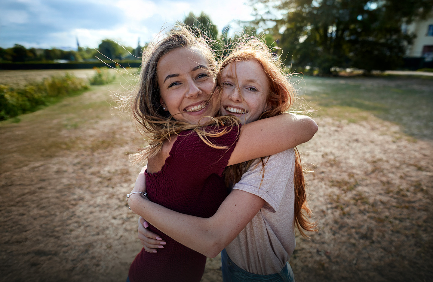 Zwei junge Frauen stehen auf eine Wiese und umarmen sich. Sie lachen beide in die Kamera.