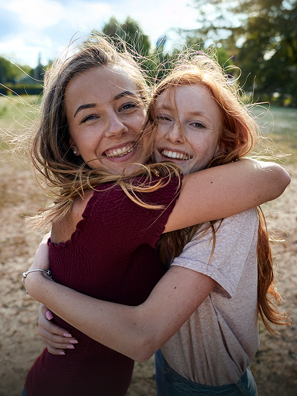 Zwei junge Frauen stehen auf eine Wiese und umarmen sich. Sie lachen beide in die Kamera.