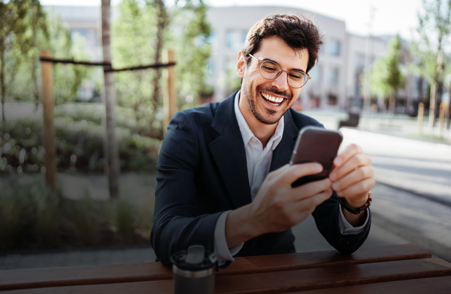 Ein Mann sitzt im Freien auf einer Bank und Blickt mit einem breiten Lächeln auf sein Mobiltelefon.